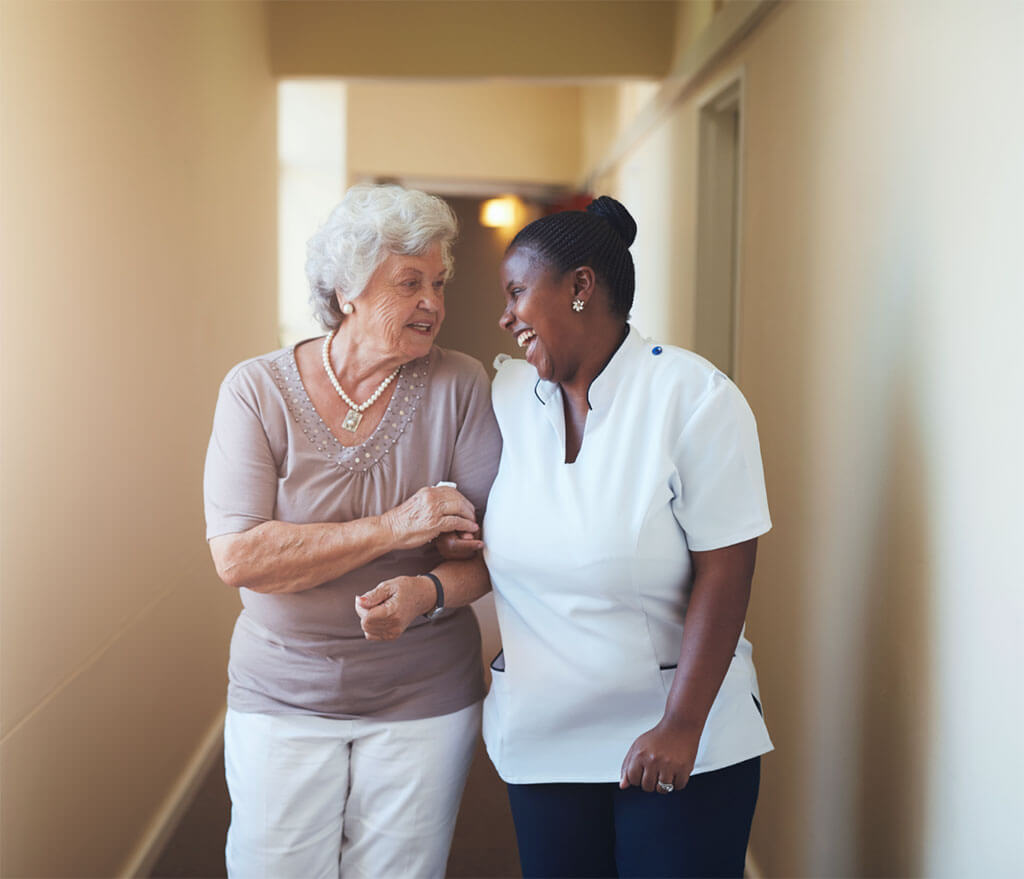 Home healthcare worker helps elderly woman walk down hallway.